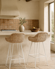 Pair of woven rattan barstools with white metal legs styled at a Mediterranean-inspired rendered kitchen island with natural light, rustic tiled splashback, and exposed timber beams.