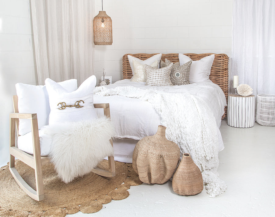 Serene bedroom featuring a rattan bedhead styled with white linen bedding, textured neutral-toned cushions, and woven decor. A natural timber rocking chair with plush cushions and a soft sheepskin throw sits on a round jute rug, complementing the airy, coastal aesthetic