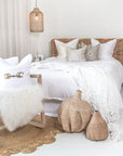Serene bedroom featuring a rattan bedhead styled with white linen bedding, textured neutral-toned cushions, and woven decor. A natural timber rocking chair with plush cushions and a soft sheepskin throw sits on a round jute rug, complementing the airy, coastal aesthetic