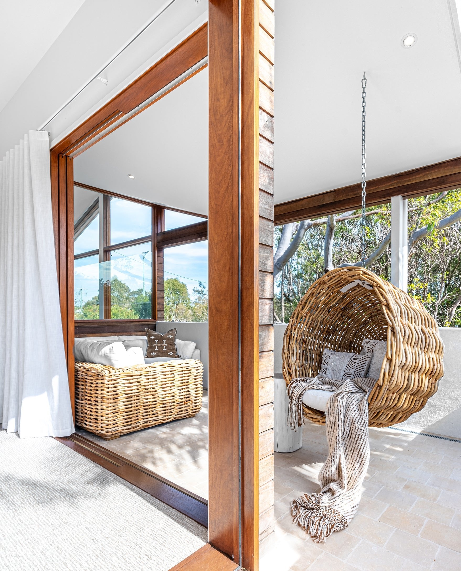 Natural wicker hanging egg chair with white cushion and throw, styled in a white outdoor patio space with built-in seating, firewood storage, and a pizza oven