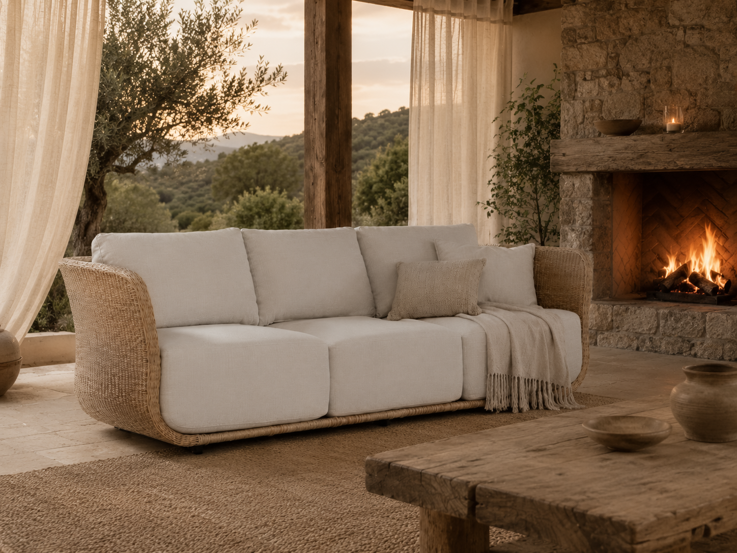 Malusi Sofa with a woven rattan frame and natural upholstered cushions styled in a warm resort-inspired living room beside a stone fireplace with flowing linen curtains and countryside views