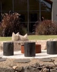A serene outdoor setting featuring KULALA Living’s black wooden stools arranged around a rusted steel firepit. Set against a modern black-clad home with lush greenery, this space blends contemporary design with natural elements for a stylish retreat. Neutral linen cushions add comfort to the inviting ambiance