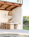 Outdoor dining setting featuring Uniqwa Collections teak chairs and travertine-look dining table beside a pool under a woven pendant light