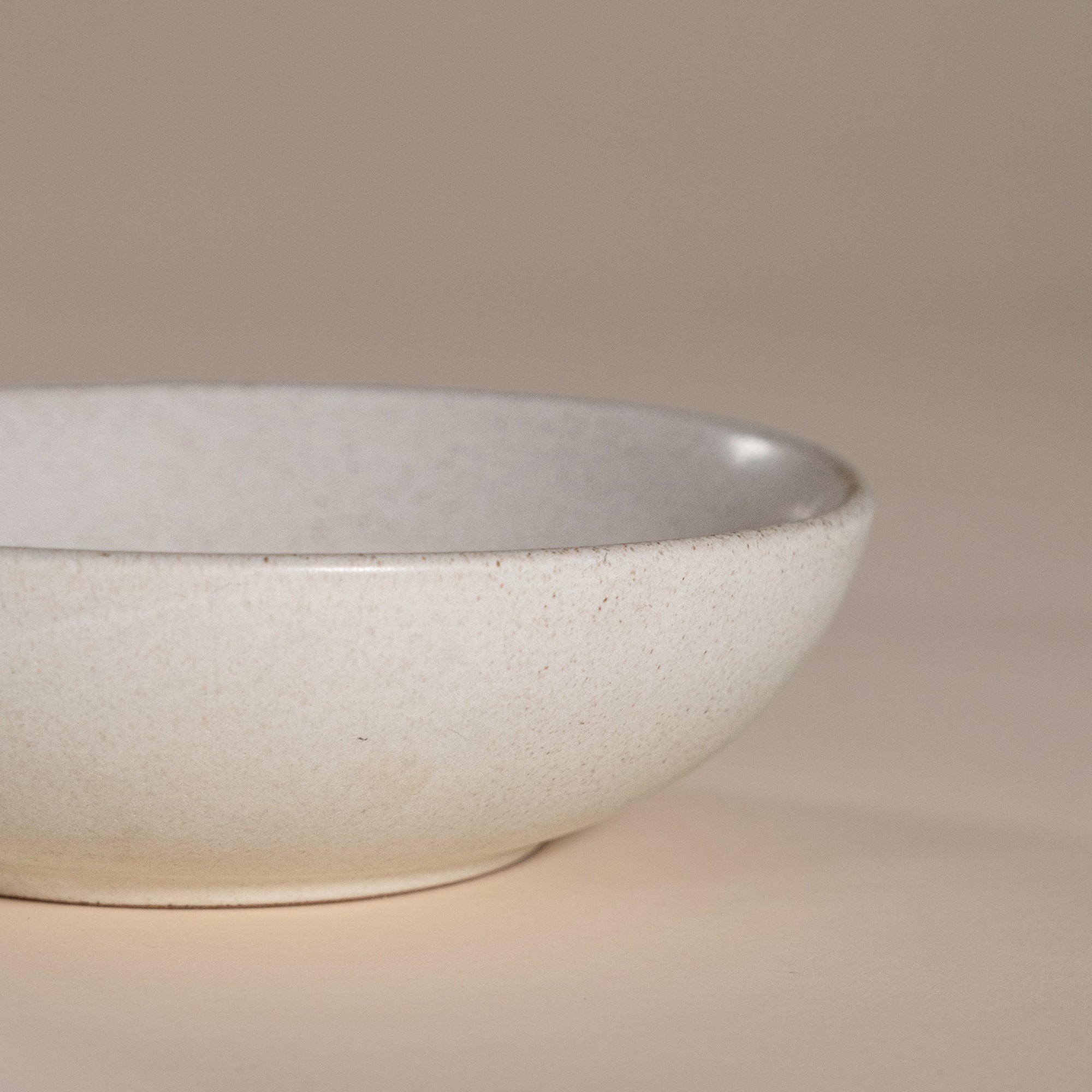 A handmade ceramic bowl with a smooth, speckled neutral glaze, set against a soft beige background. The minimalist design and organic shape make it a versatile piece for modern dining and home decor