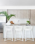 A bright modern kitchen featuring Kulala Living Makena Bar Chairs in crisp white, offering a sculptural rattan backrest and plush seating. Styled with neutral tones, natural wood cabinetry, and fresh greenery for an inviting, coastal-inspired space
