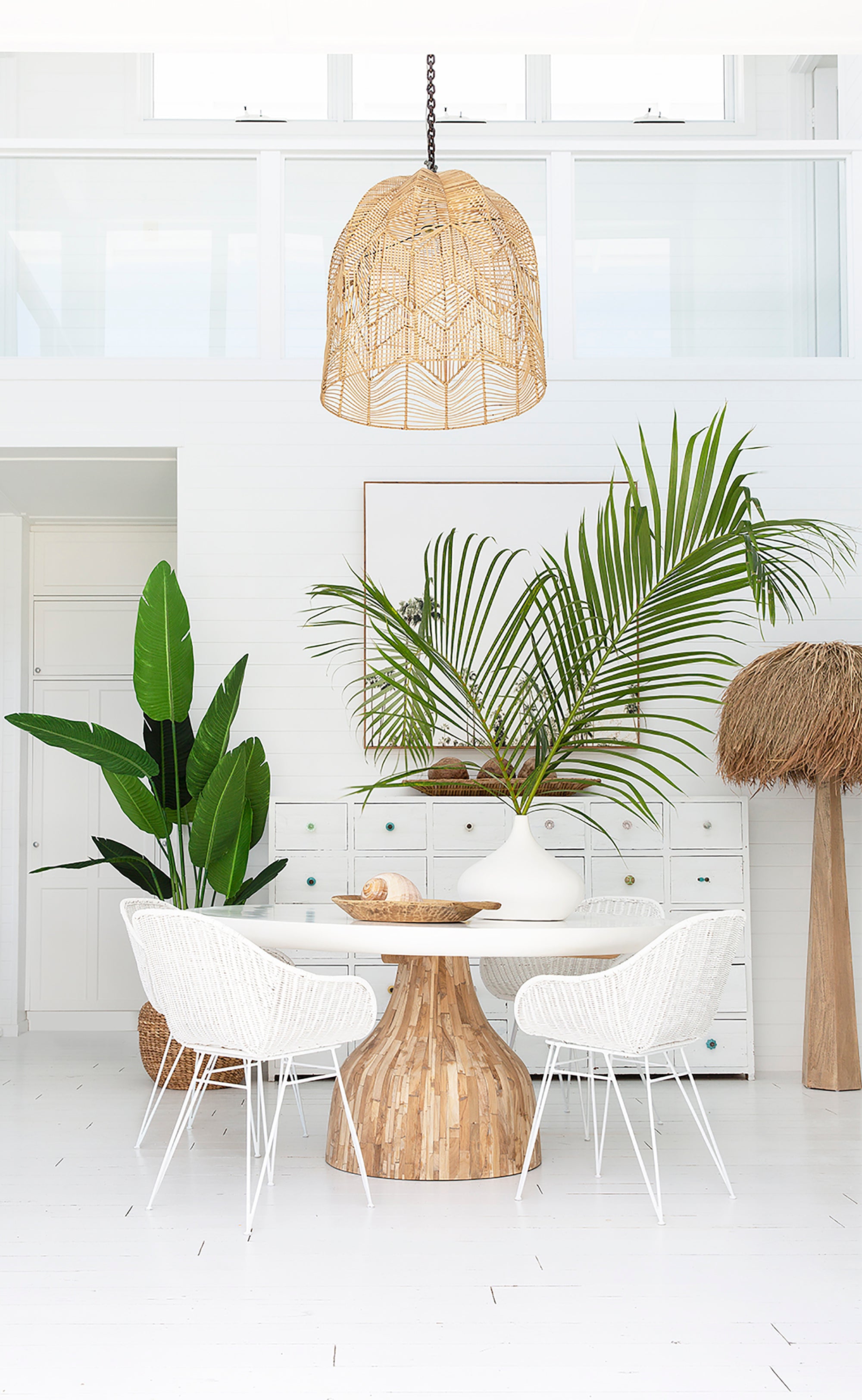Bright coastal dining setting with white rattan dining chairs, natural wood round table, and woven pendant light. Styled with lush green plants and natural decor accents