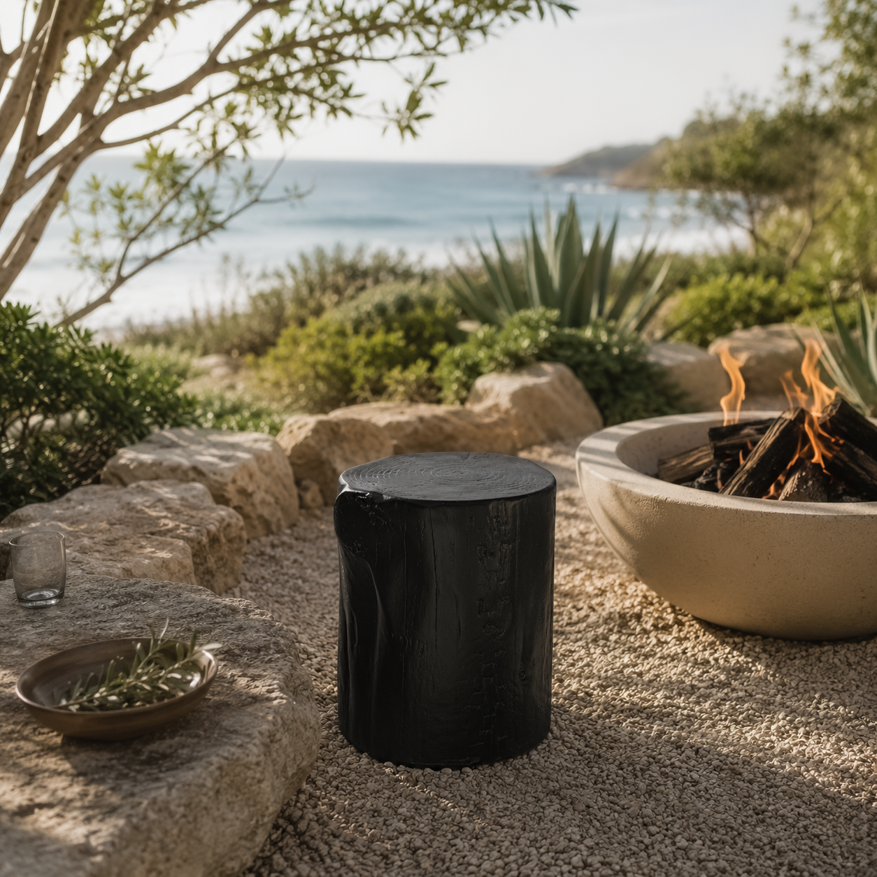 Black magnesium oxide stools arranged around an outdoor fire pit in a coastal garden retreat, creating a relaxed gathering space framed by native greenery and ocean views.