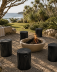 Black magnesium oxide stools arranged around an outdoor fire pit in a coastal garden retreat, creating a relaxed gathering space framed by native greenery and ocean views.