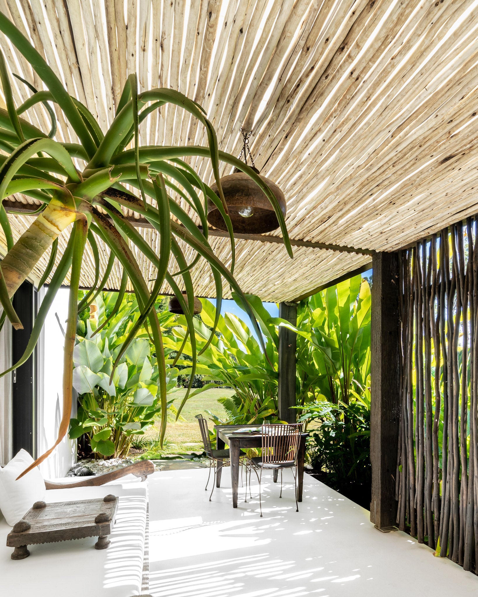 Tropical outdoor dining area under a eucalyptus pole pergola, featuring lush greenery, a rustic wooden dining table with metal chairs, and hanging pendant lighting. The space exudes a relaxed, resort-style ambiance