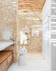 Tranquil outdoor shower with natural stone walls, brass fixtures, timber slatted ceiling, and white rendered details, styled with soft towels and earthy decor for a day spa atmosphere.