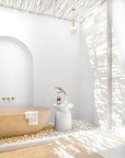 Tranquil outdoor bathroom with a sculptural stone bathtub surrounded by smooth white pebbles, minimalist white side table, and natural eucalyptus pole roof casting dappled light.