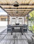 Outdoor dining area featuring a black timber dining table and woven chairs under a rustic eucalyptus pergola, with handcrafted pendant lights and lush tropical surroundings.