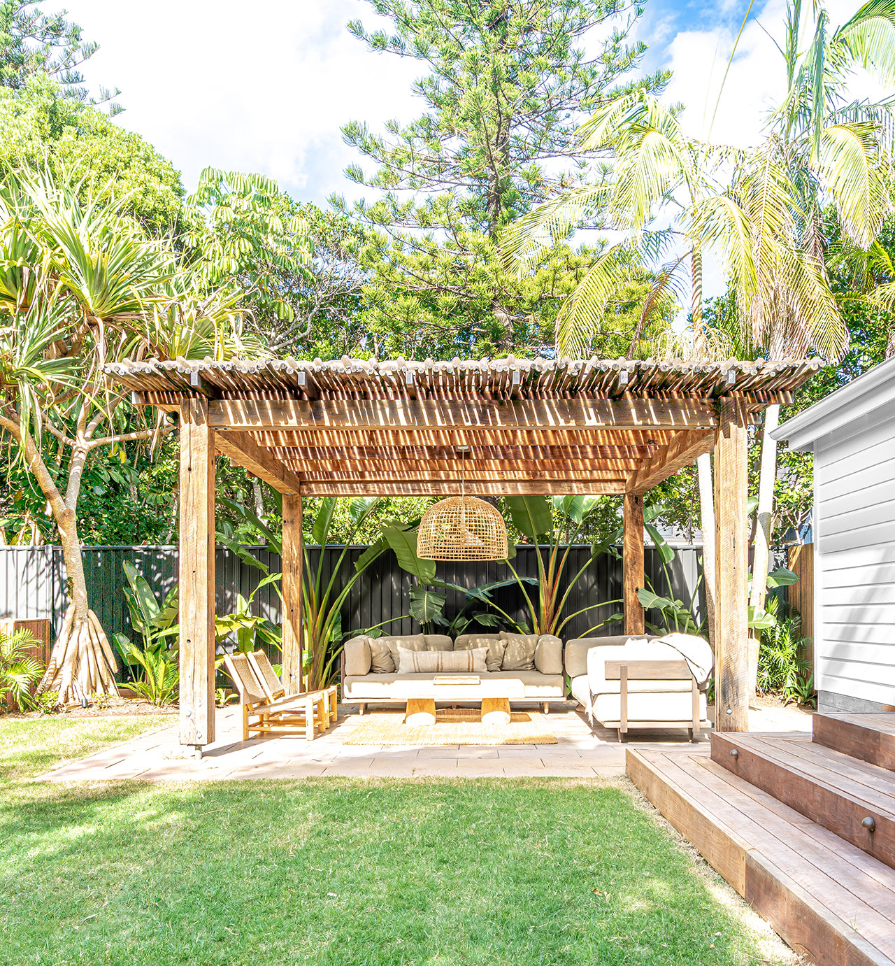 Tranquil outdoor pavilion featuring a natural timber pergola, latte pole roof, rattan pendant light, teak outdoor sofa, and tropical garden setting with lush greenery.