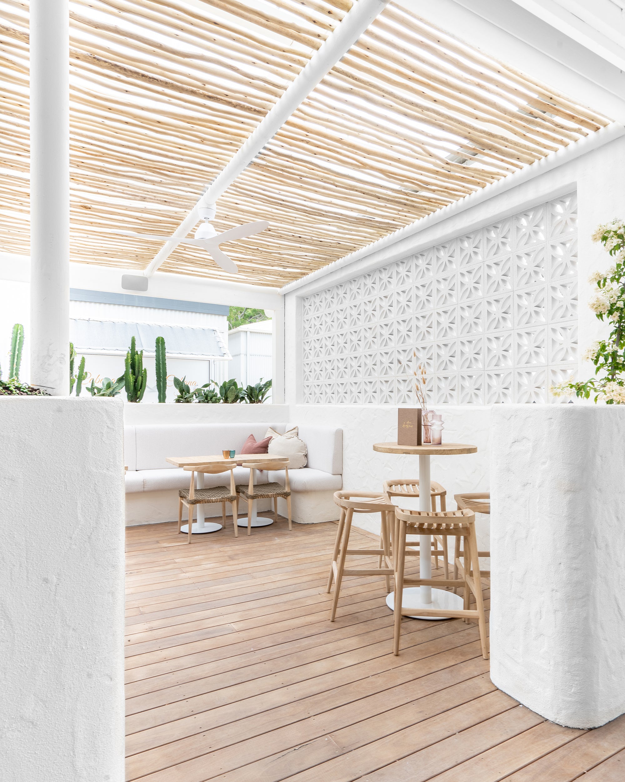 Light-filled courtyard space with white breeze block feature wall, rattan stools, timber dining tables, and white banquette seating surrounded by greenery and natural textures.